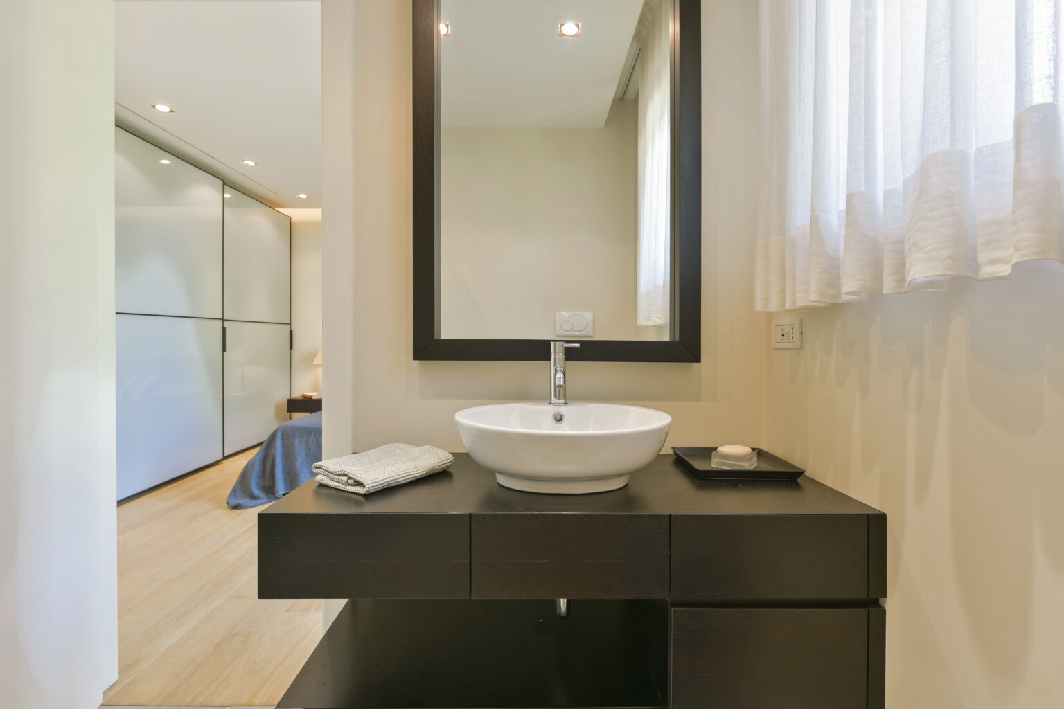 Modern bathroom interior featuring a sleek black vanity with a round white sink, a large mirror, and natural light from a nearby window. The room is designed with minimalist decor, showcasing light wood flooring and a contemporary aesthetic. | Only on SopranoVillas