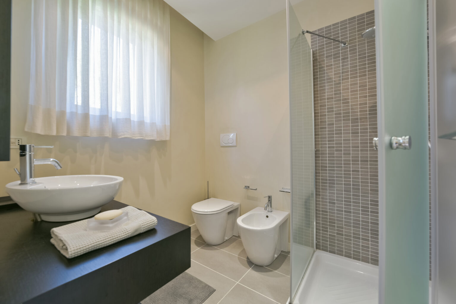 Modern bathroom featuring a stylish shower, bidet, and elegant sink with a countertop, complemented by natural light from a window and neutral wall colors. | Only on SopranoVillas