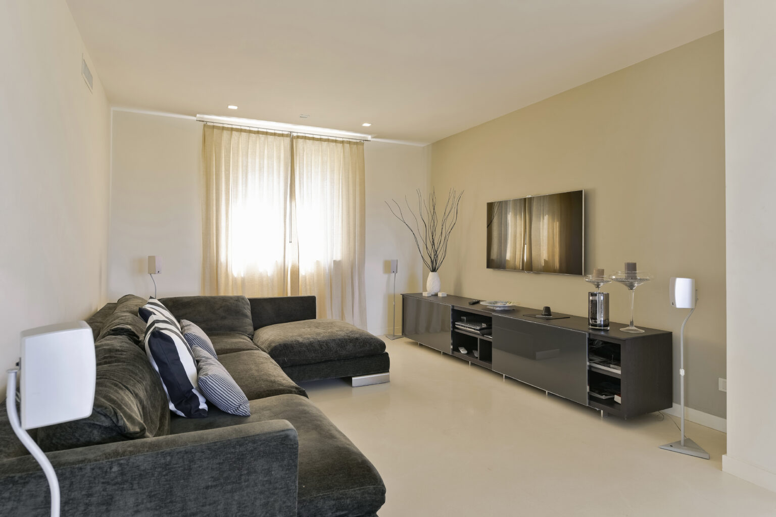 Modern living room featuring a black sectional sofa, light-colored walls, and large windows with sheer curtains. A flat-screen TV is mounted on a sleek entertainment unit, complemented by decorative elements like a floor lamp and a vase. The space is bright and inviting, showcasing contemporary interior design. | Only on SopranoVillas