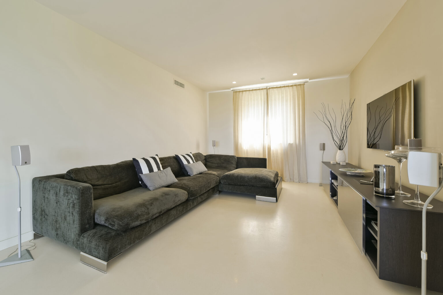 Modern living room featuring a large dark green sectional sofa with decorative pillows, a sleek entertainment unit, and large windows with sheer curtains allowing natural light to fill the space. | Only on SopranoVillas