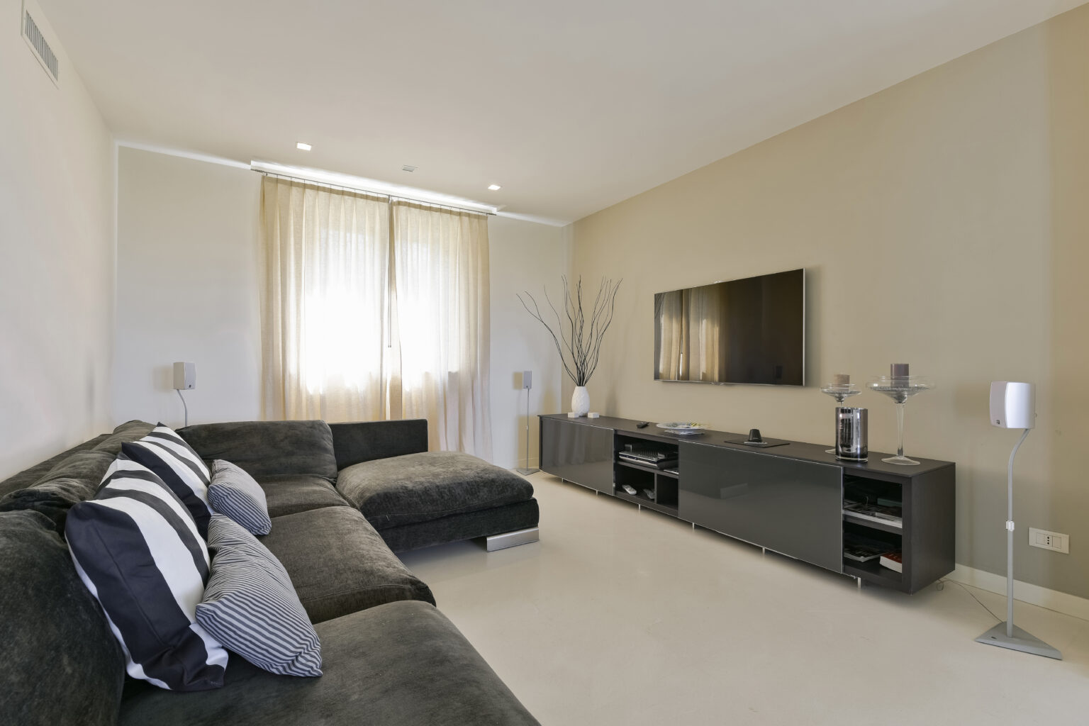 Modern living room featuring a dark sectional sofa with decorative pillows, a sleek entertainment unit with a flat-screen TV, and large windows draped with light curtains, creating a bright and inviting atmosphere. | Only on SopranoVillas
