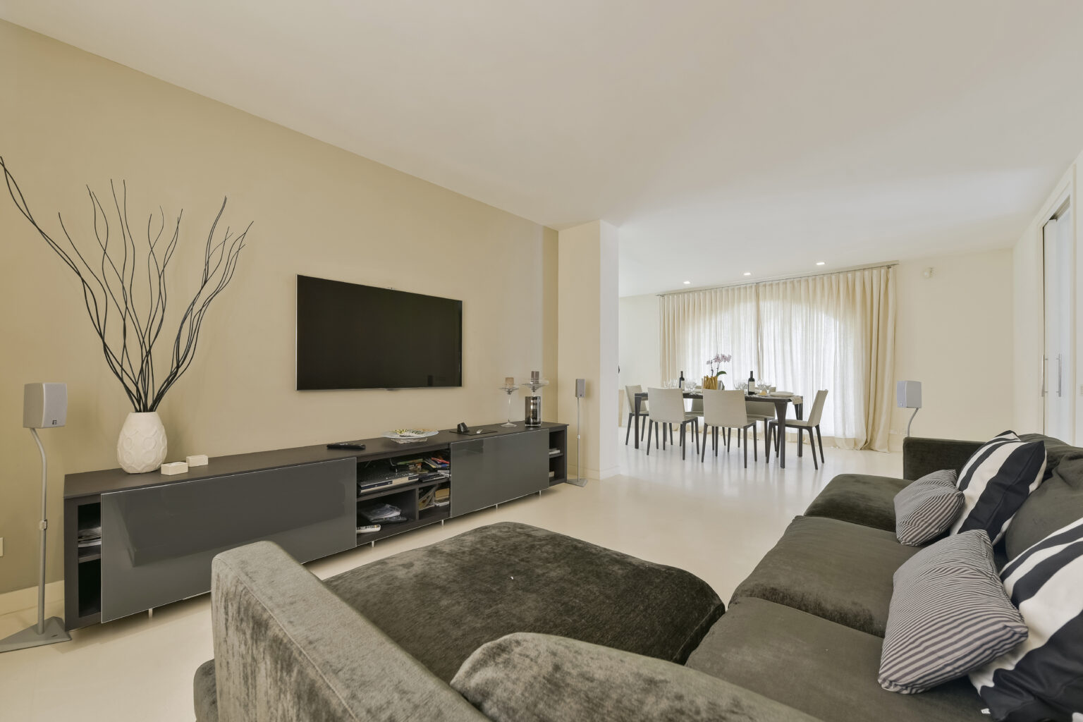 Modern living room interior featuring a comfortable grey sofa, a sleek entertainment unit with a flat-screen TV, and a dining area visible in the background. The space is adorned with minimalist decor, including decorative branches in a vase and soft lighting from large windows. | Only on SopranoVillas