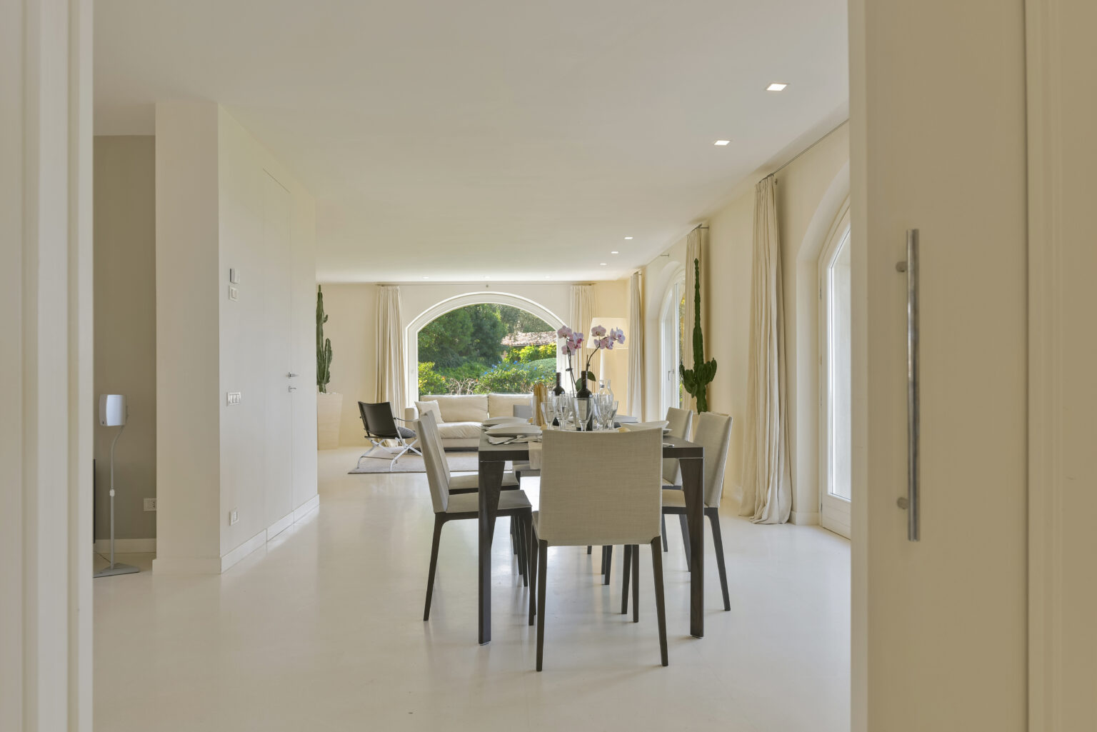 Modern dining area with a sleek table and chairs, featuring large windows that allow natural light and a view of the garden. The space is elegantly decorated with neutral tones and soft textures, creating a warm and inviting atmosphere. | Only on SopranoVillas