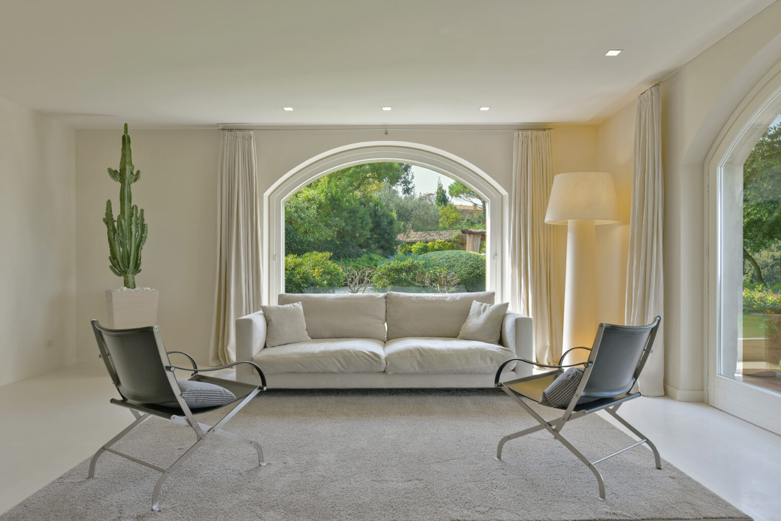 Modern living room featuring a light-colored sofa, two sleek chairs, and large arched windows with views of greenery. The space is illuminated by natural light and accented with a tall cactus in a decorative pot. | Only on SopranoVillas