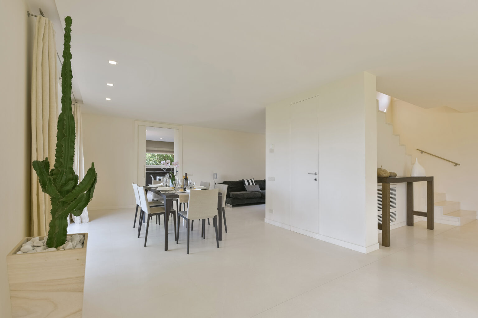 Modern dining area with a sleek table and chairs, complemented by a stylish cactus plant in a planter. The bright, open space features neutral walls and a staircase, creating an inviting atmosphere. Ideal for contemporary home design inspiration. | Only on SopranoVillas