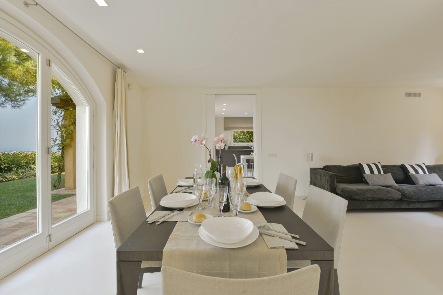 Modern dining area featuring a stylish table set for a meal, with elegant tableware and floral centerpiece. Bright natural light streams through large windows, highlighting the open layout that connects to a cozy living room with a dark sofa. | Only on SopranoVillas