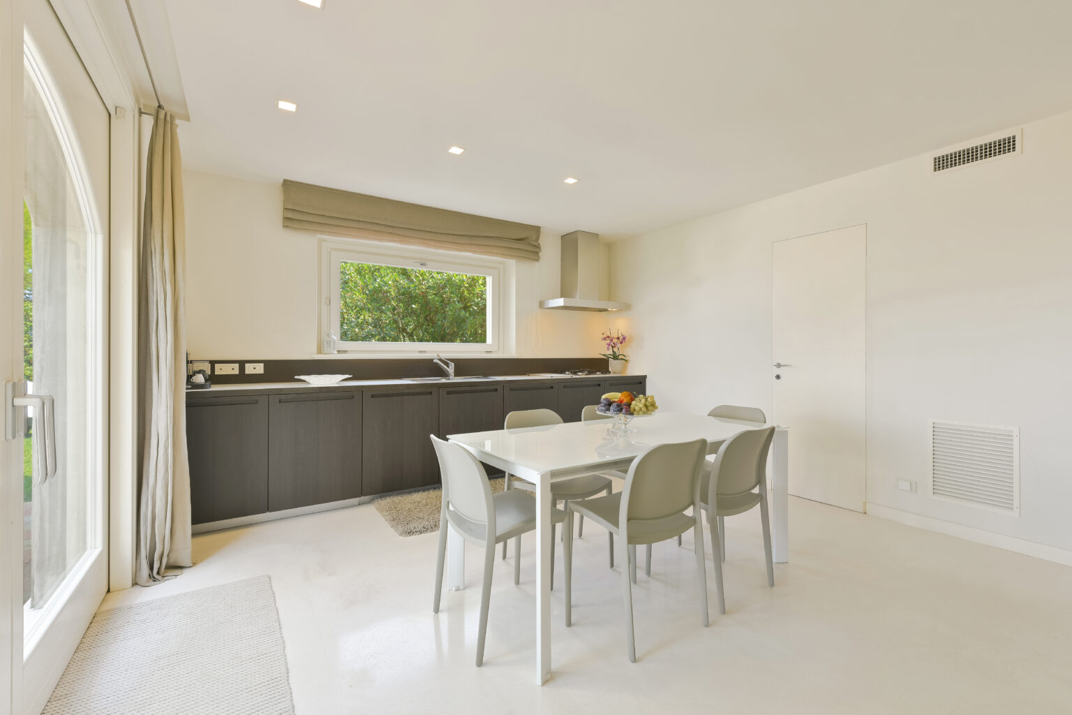 Modern kitchen and dining area featuring a sleek black cabinetry, white walls, and a dining table with four chairs. Natural light floods the space through large windows, creating a bright and inviting atmosphere. | Only on SopranoVillas