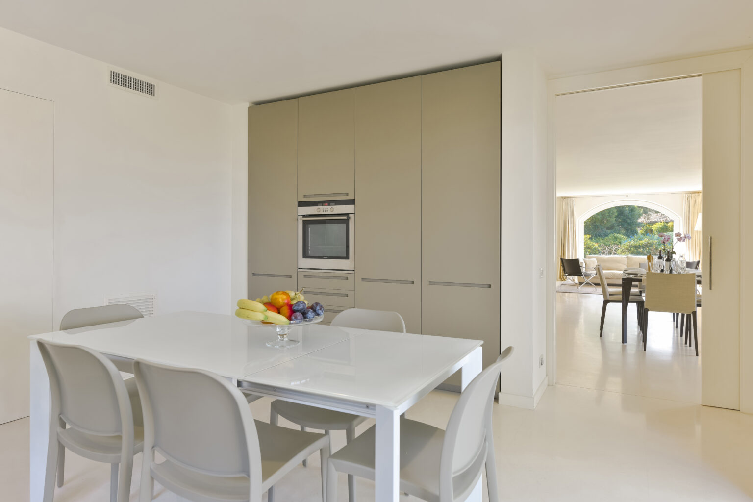 Modern kitchen interior featuring a sleek white dining table with fruit centerpiece, minimalist cabinetry, and an open layout leading to a bright living space. | Only on SopranoVillas