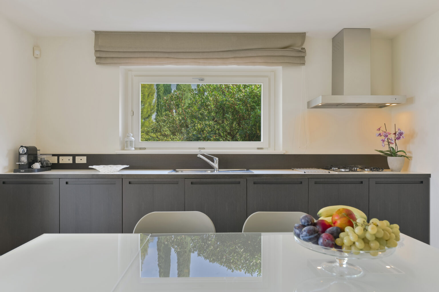 Modern kitchen interior featuring sleek black cabinetry, a white countertop, and a window with a view of greenery. A bowl of fresh fruit, including grapes and figs, is displayed on the table, adding a touch of color to the space. | Only on SopranoVillas