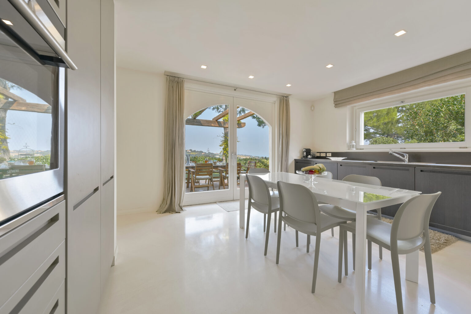 Modern kitchen interior with sleek cabinetry, white dining table, and chairs, featuring large windows that open to a scenic outdoor view. Bright, airy atmosphere enhanced by natural light. Ideal for contemporary home design inspiration. | Only on SopranoVillas