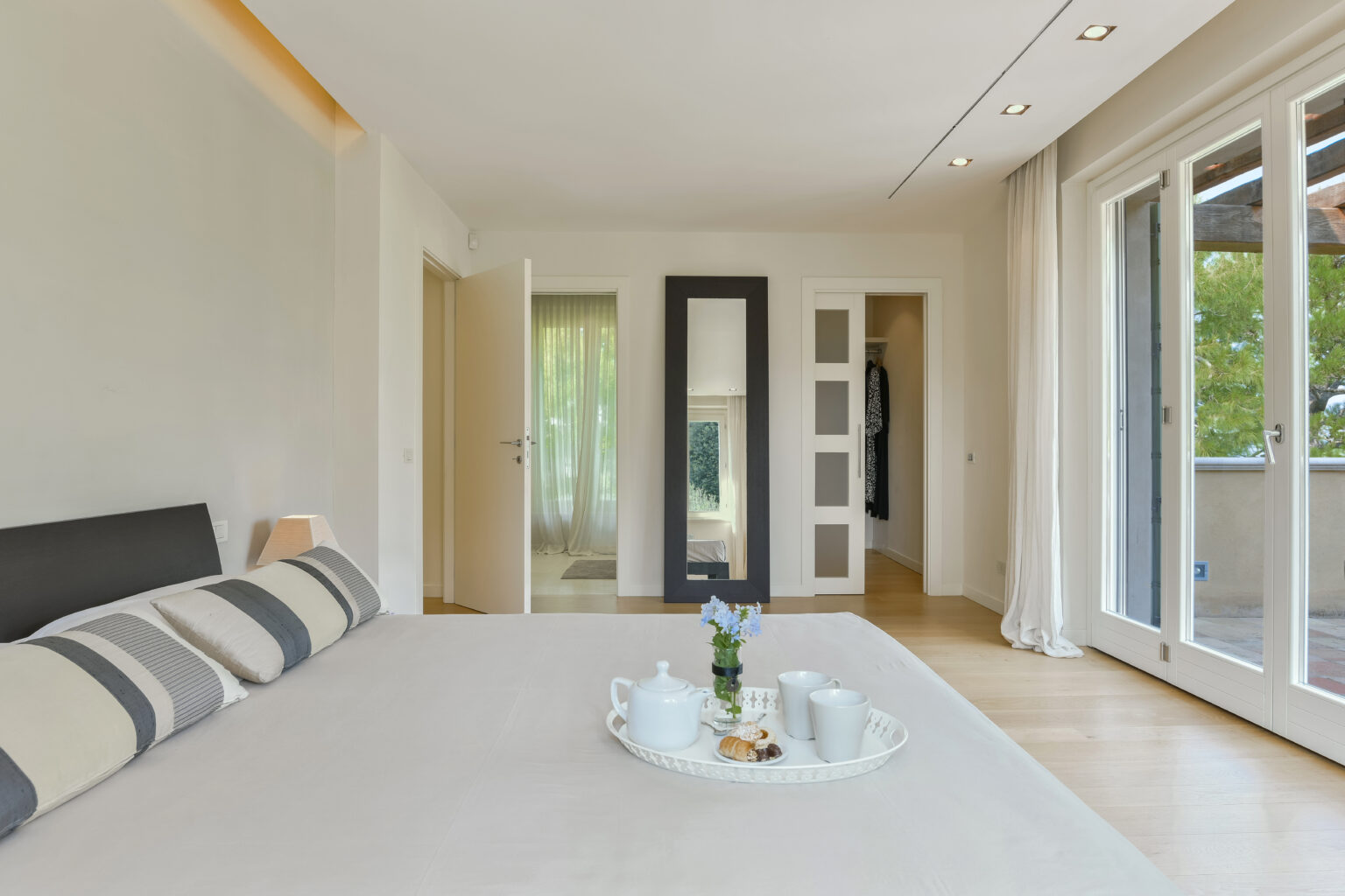 Modern bedroom with a minimalistic design featuring a bed with striped pillows, a decorative tray with tea set, and large windows that open to a balcony, allowing natural light to fill the space. | Only on SopranoVillas
