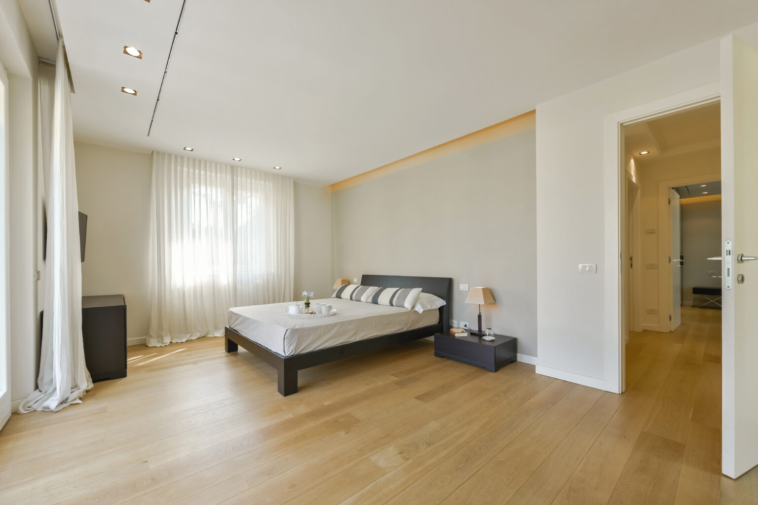 Modern bedroom interior featuring a queen-sized bed with decorative pillows, wooden flooring, large windows with sheer curtains, and minimalist furniture including nightstands and lamps. | Only on SopranoVillas