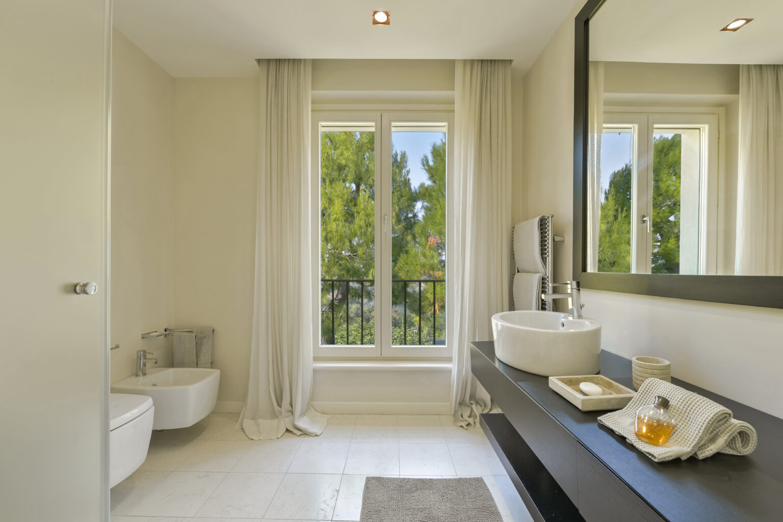 Modern bathroom interior featuring a sleek design, large window with a view of greenery, minimalist sink, and elegant decor with towels and a tea set on a dark wood countertop. | Only on SopranoVillas