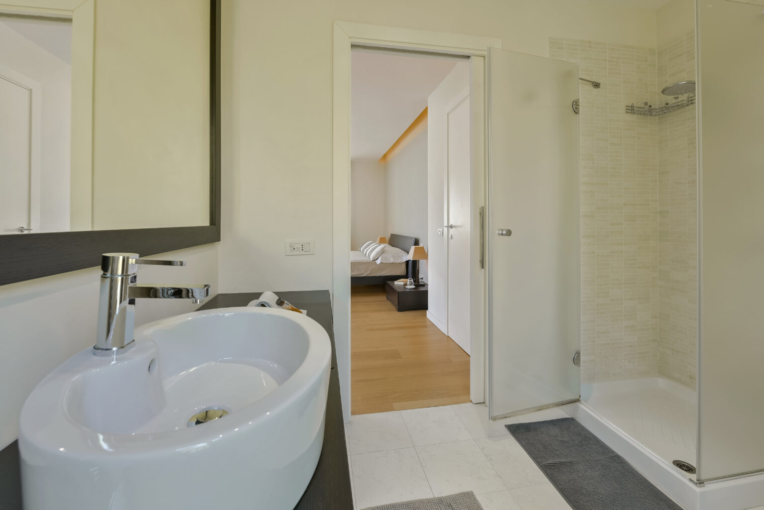 Modern bathroom featuring a sleek sink and shower, with a view into a well-lit bedroom. The design emphasizes clean lines and a minimalist aesthetic, showcasing a spacious and inviting atmosphere. | Only on SopranoVillas