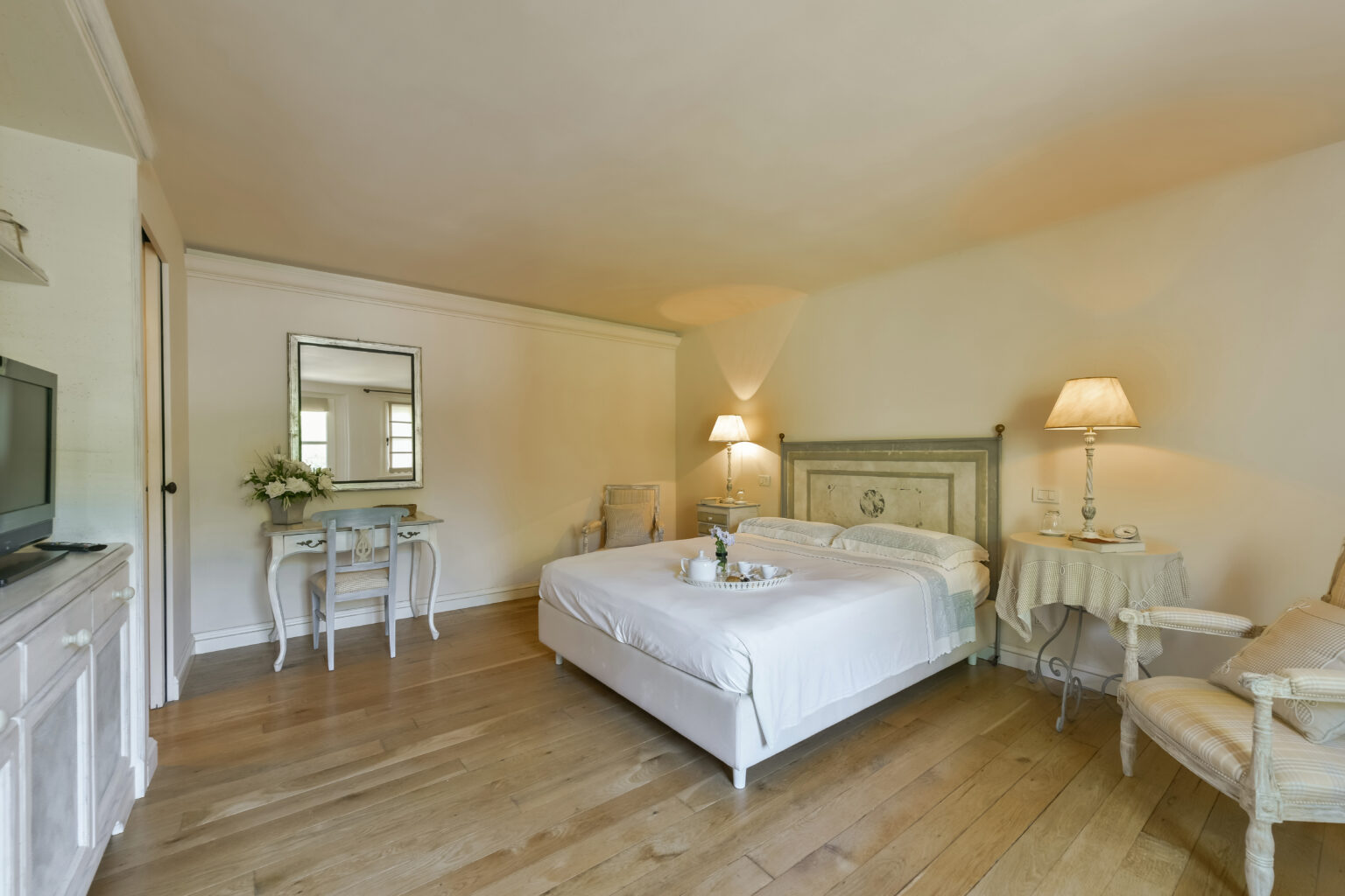 Cozy and elegantly designed bedroom featuring a comfortable double bed, wooden flooring, soft lighting from bedside lamps, and a stylish seating area. The room includes a mirror and a small table, creating a welcoming atmosphere for relaxation. Ideal for a serene getaway or a restful night. | Only on SopranoVillas