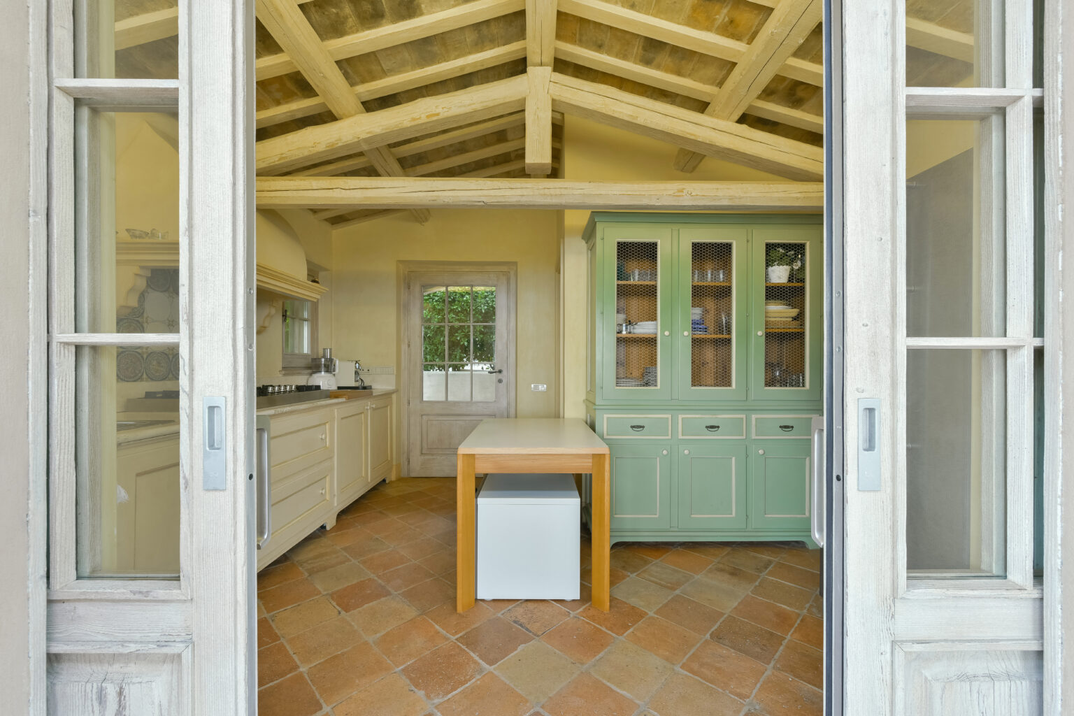 Bright and airy kitchen with rustic wooden beams, featuring white cabinetry, a green hutch, and a central wooden island, viewed through double doors. | Only on SopranoVillas