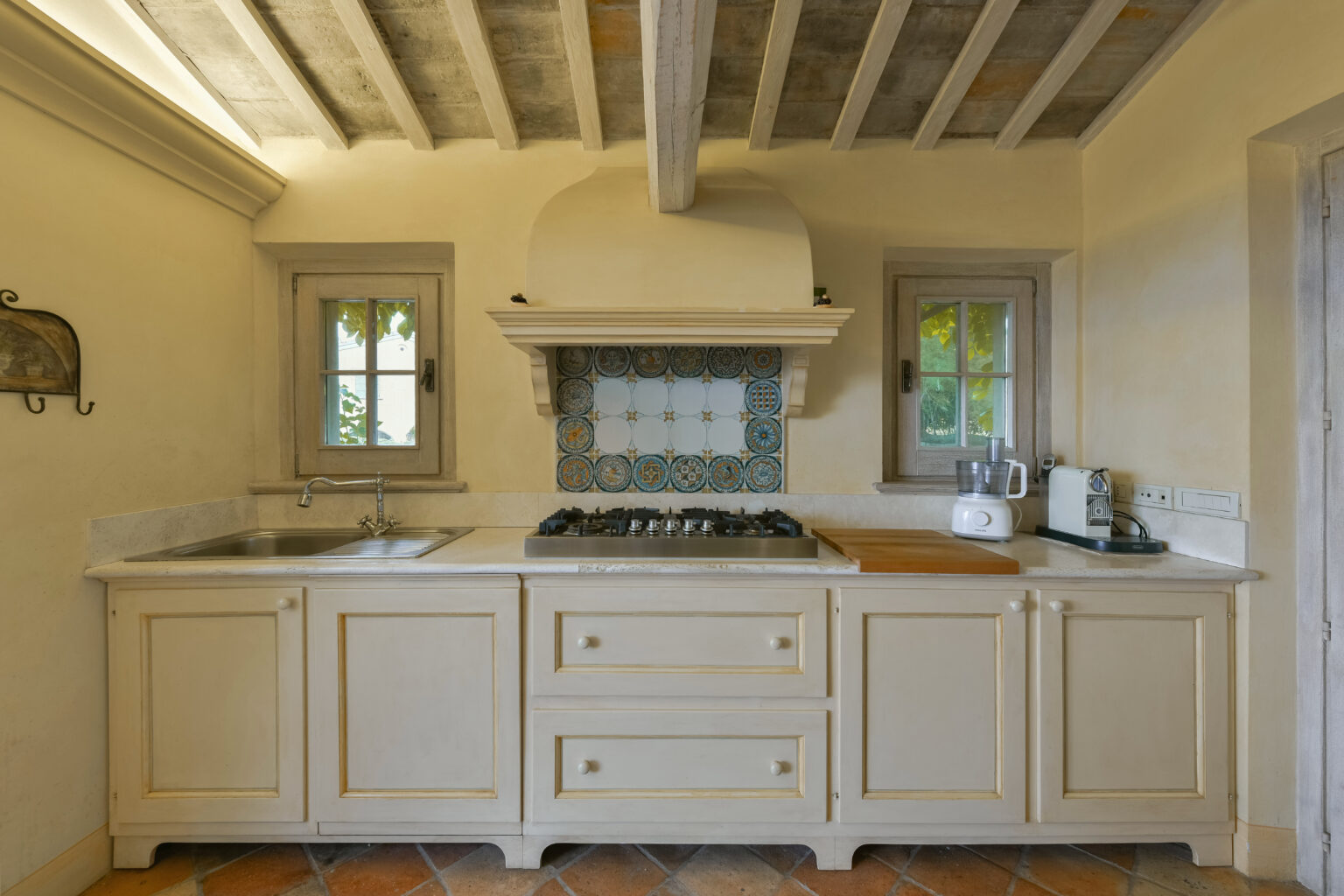 Charming rustic kitchen with cream-colored cabinetry, a gas stove, and tiled backsplash, featuring natural light from small windows and wooden ceiling beams. | Only on SopranoVillas
