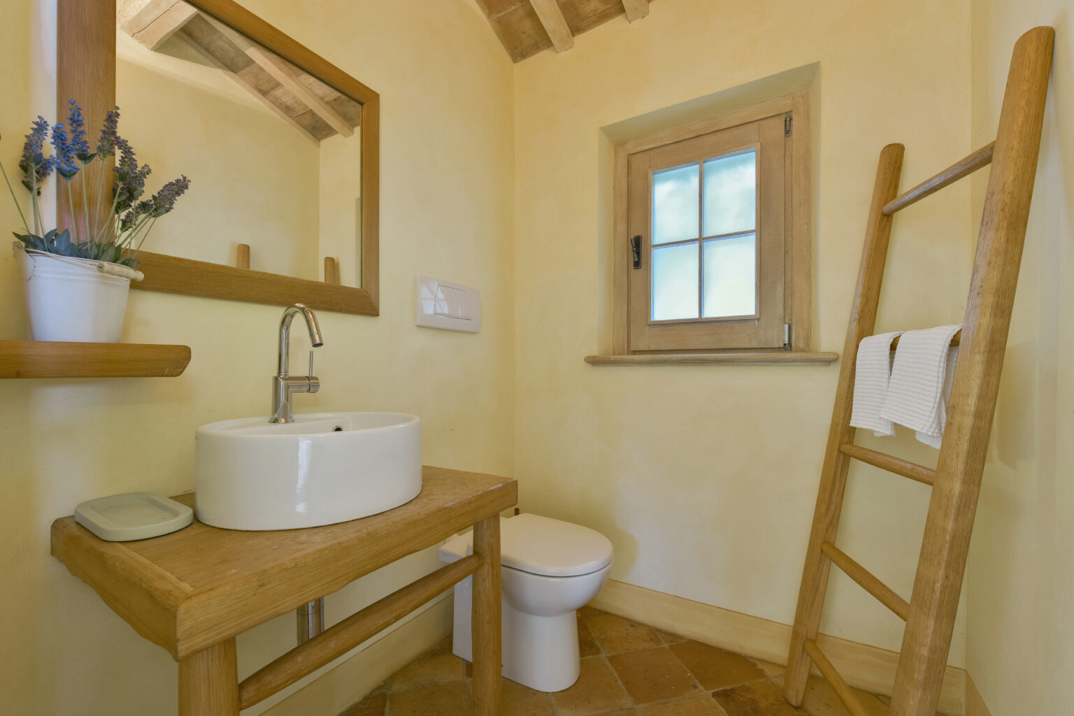 Modern bathroom with a wooden sink table, white vessel sink, and a toilet, featuring a rustic ladder for towels and a small window with natural light. Decorated with a potted plant for a touch of greenery. | Only on SopranoVillas