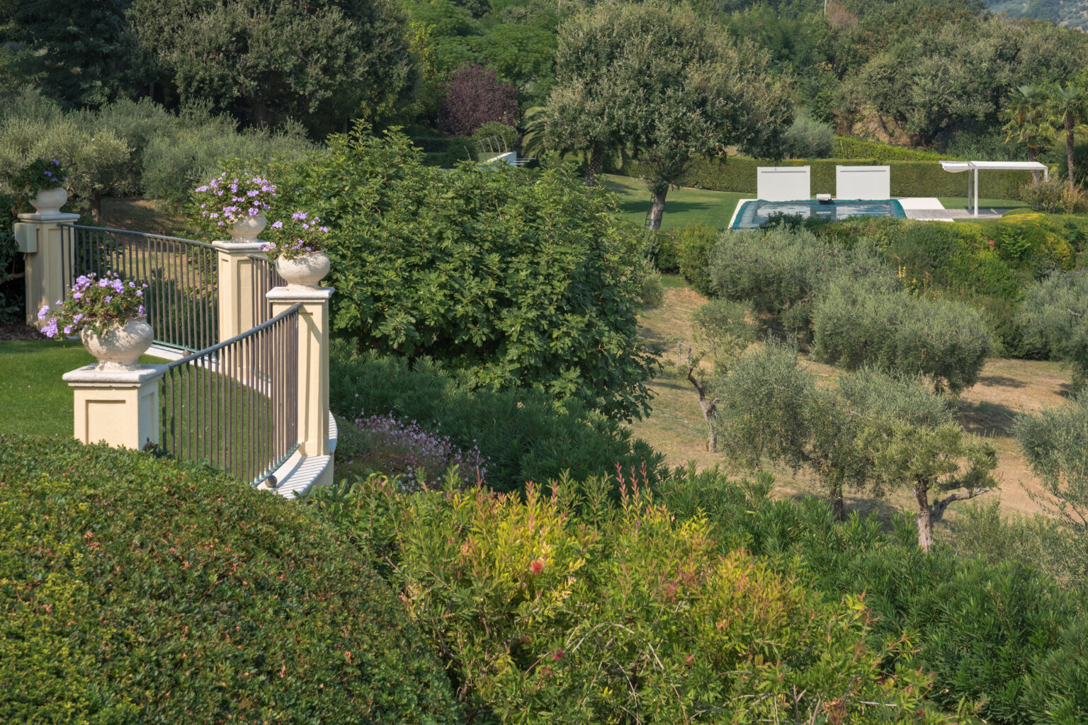 Lush green landscape featuring manicured gardens, olive trees, and a swimming pool in the background, framed by decorative stone railings and flowering plants. | Only on SopranoVillas