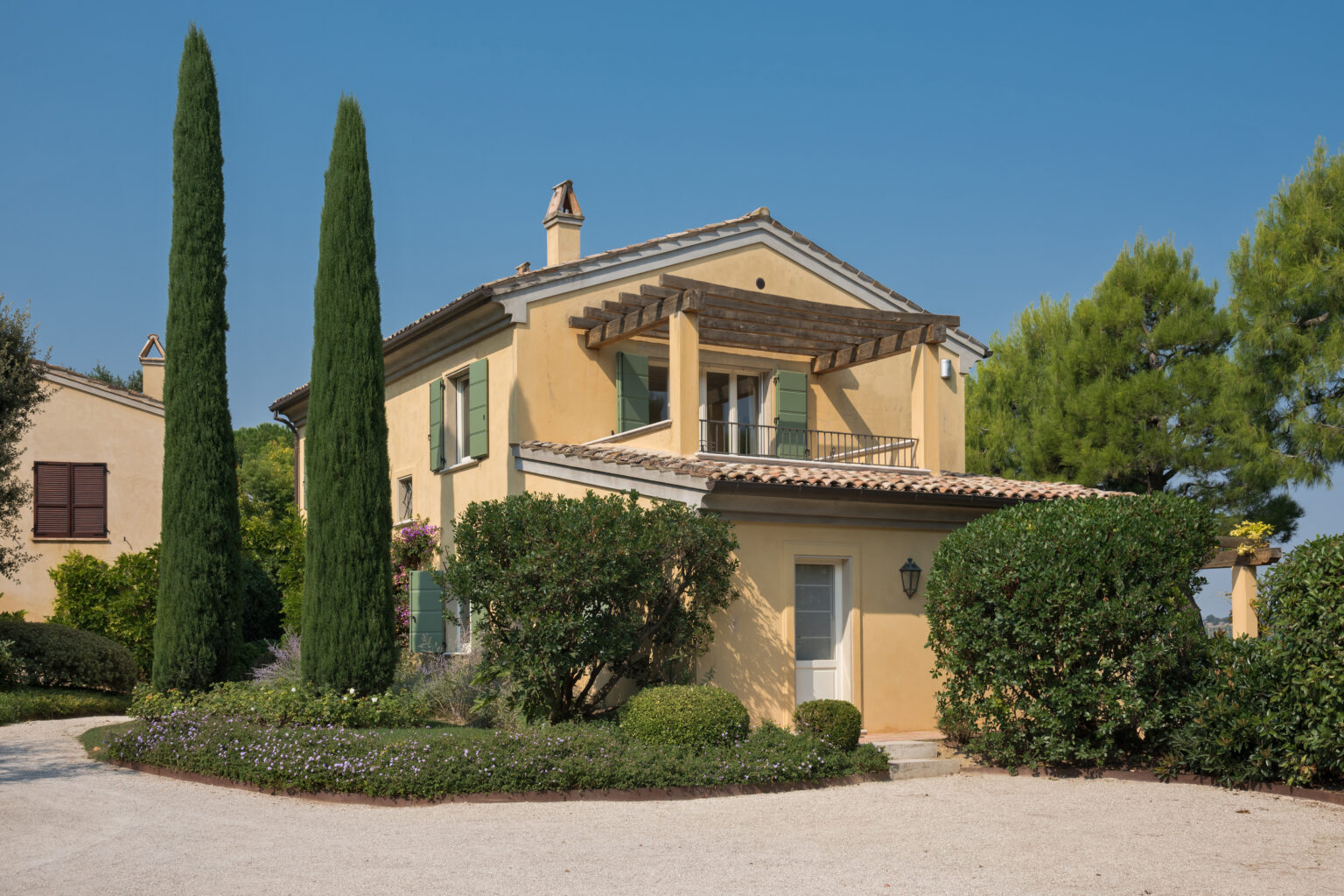 Charming yellow villa surrounded by lush greenery, featuring tall cypress trees and a well-manicured garden, set against a clear blue sky. | Only on SopranoVillas