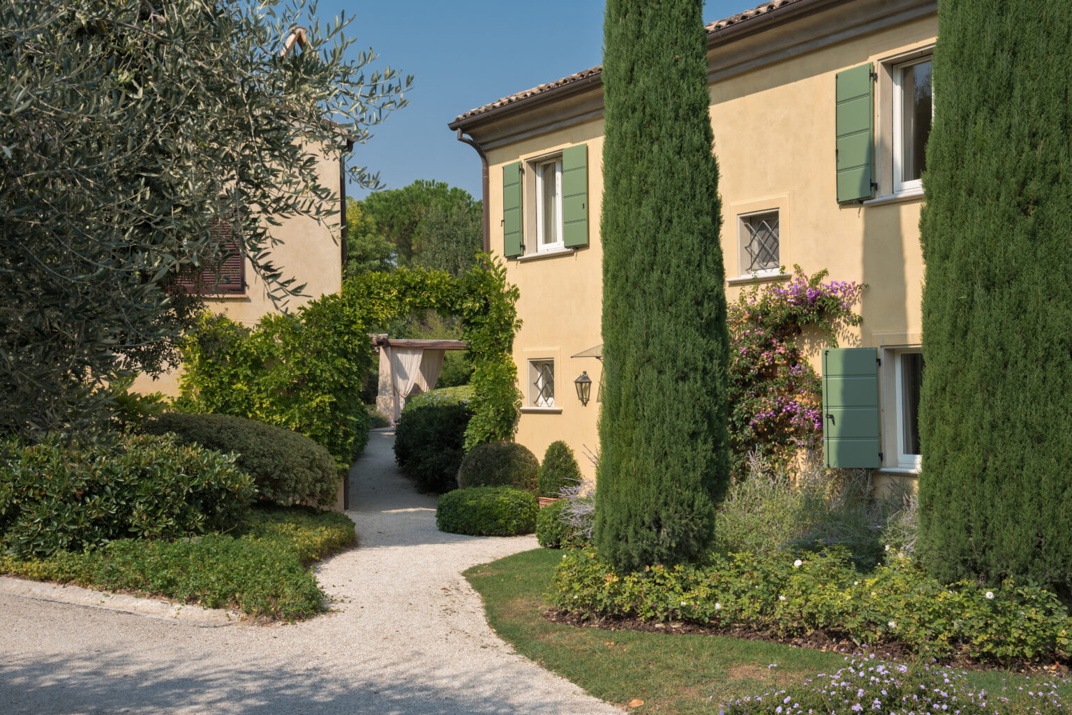 Charming residential pathway leading to a yellow house with green shutters, surrounded by lush greenery and tall cypress trees, creating a picturesque garden setting. | Only on SopranoVillas