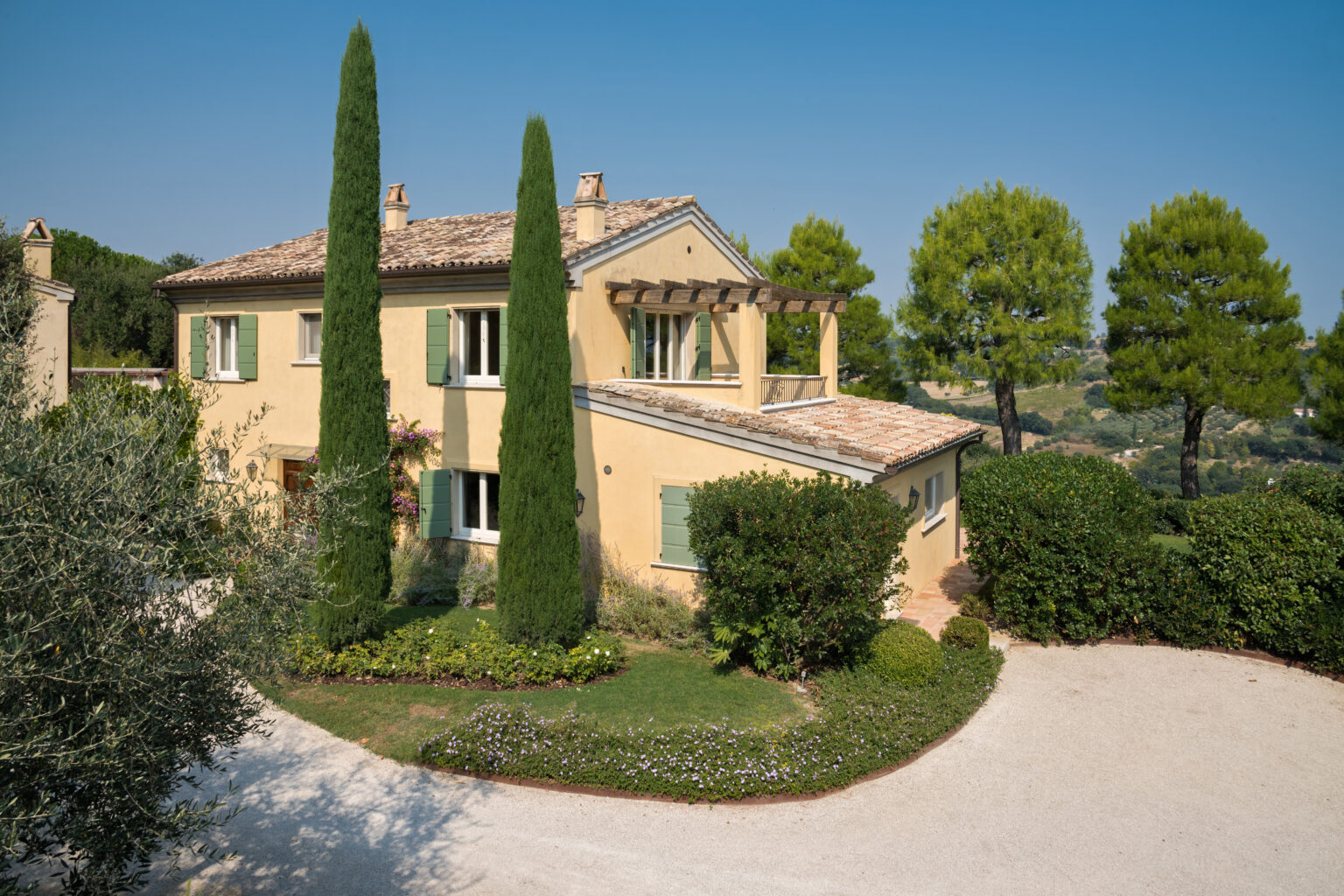 Charming yellow villa surrounded by lush greenery, featuring tall cypress trees and a gravel driveway, set against a clear blue sky. Perfect for a serene getaway or vacation rental in a picturesque landscape. | Only on SopranoVillas