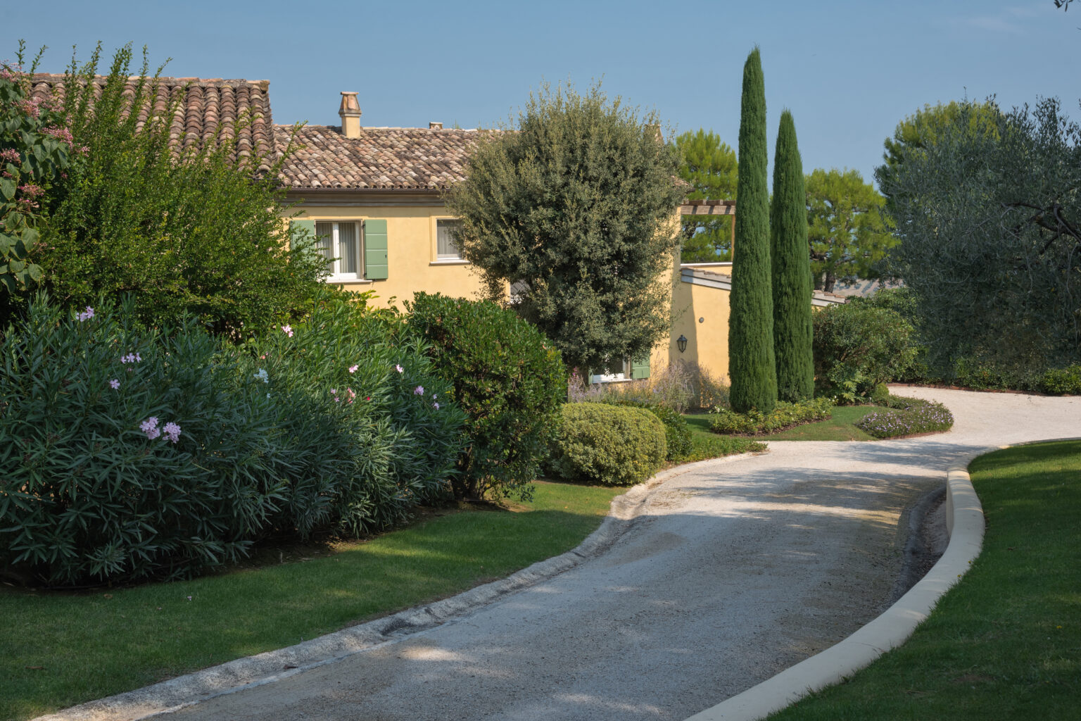 Charming countryside home with a gravel driveway surrounded by lush greenery and tall cypress trees, set against a clear blue sky. | Only on SopranoVillas