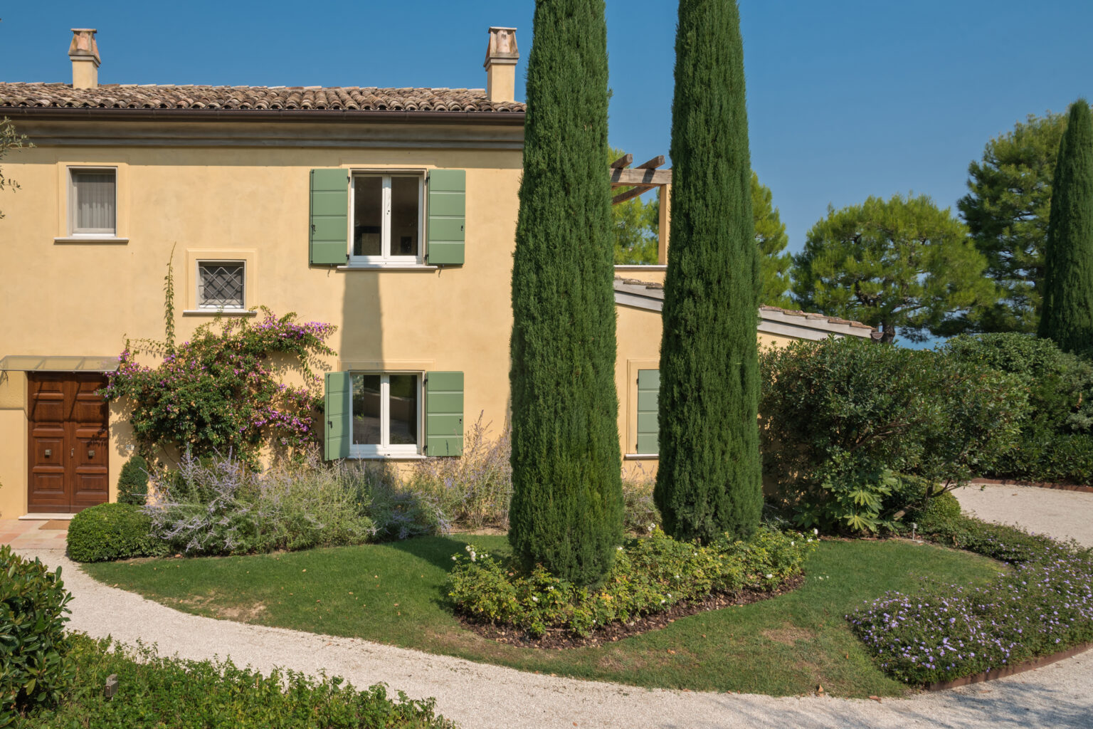 Charming yellow villa with green shutters surrounded by landscaped gardens and tall cypress trees under a clear blue sky. | Only on SopranoVillas