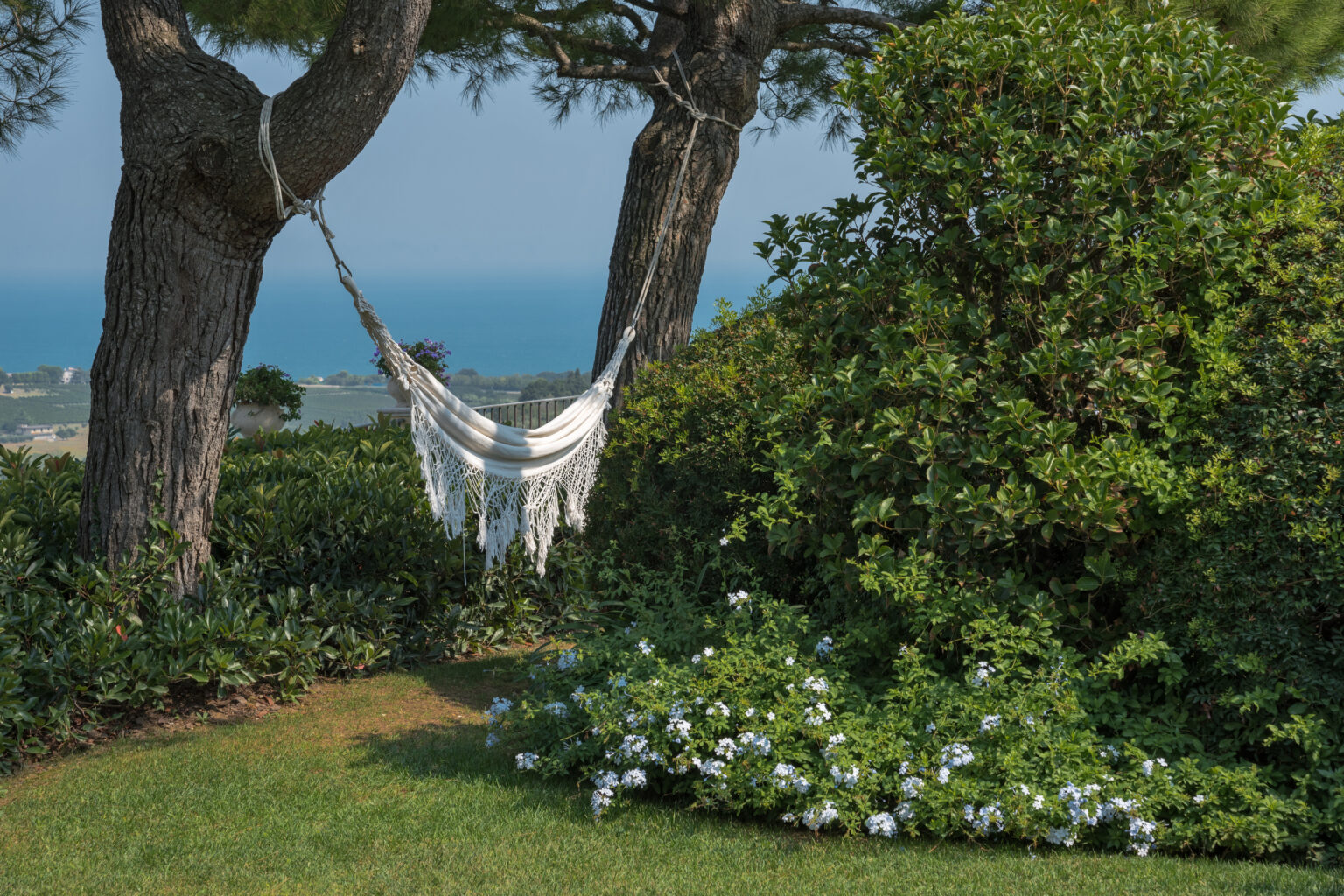 A serene outdoor scene featuring a white hammock suspended between two trees, surrounded by lush greenery and flowering plants, with a tranquil ocean view in the background. | Only on SopranoVillas