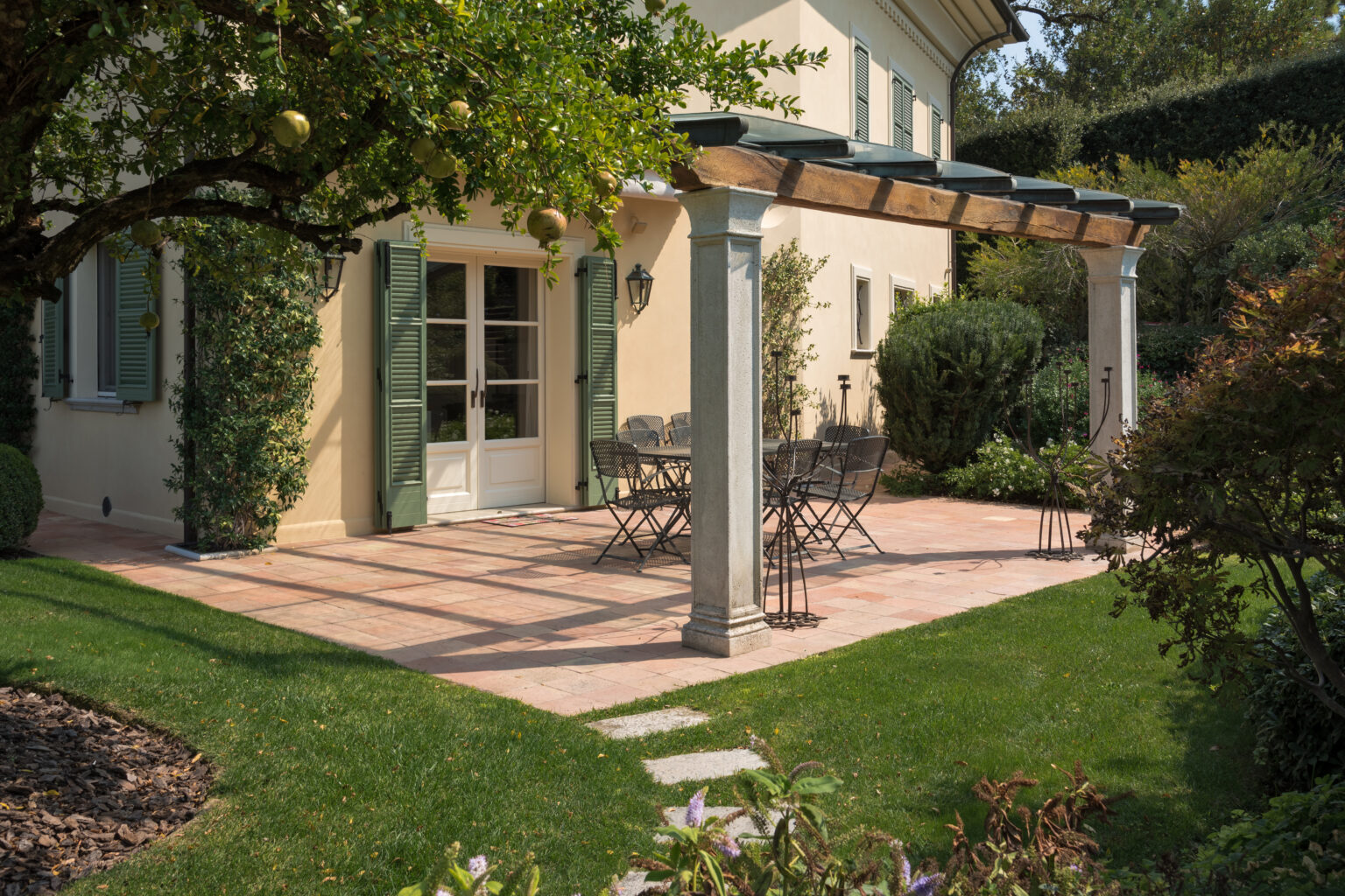 Charming outdoor patio area featuring a stone pergola, black metal dining set, and lush greenery surrounding a beige house with green shutters. Perfect for entertaining and enjoying nature. | Only on SopranoVillas