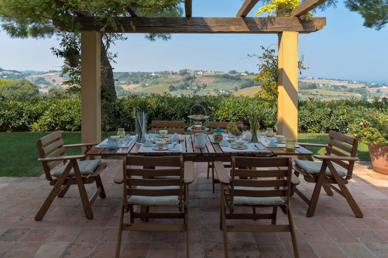 Outdoor dining table set for a meal with wooden chairs, overlooking a scenic landscape of rolling hills and greenery. | Only on SopranoVillas