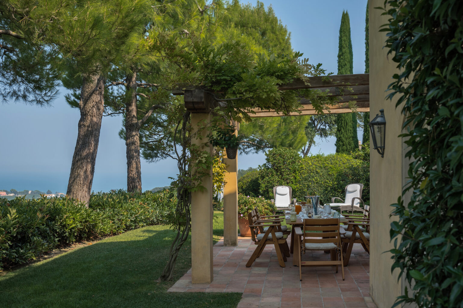 Outdoor dining area with wooden table and chairs under a pergola, surrounded by lush greenery and tall trees, overlooking a scenic view. Ideal for al fresco gatherings and relaxation. | Only on SopranoVillas
