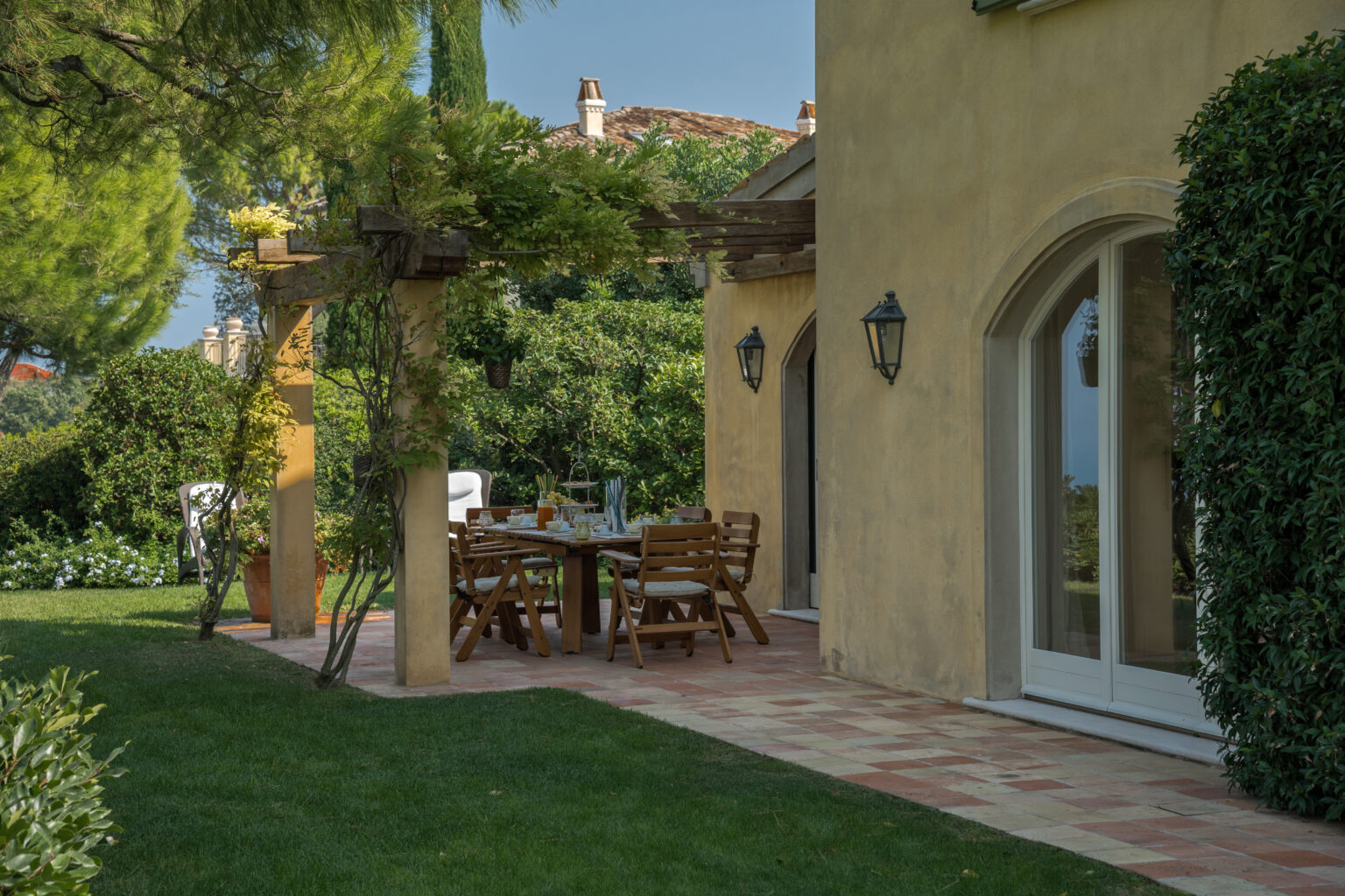 A charming outdoor dining area featuring a wooden table and chairs under a pergola, surrounded by lush greenery and a well-maintained garden, with a view of a sunny landscape and a cozy home exterior. | Only on SopranoVillas