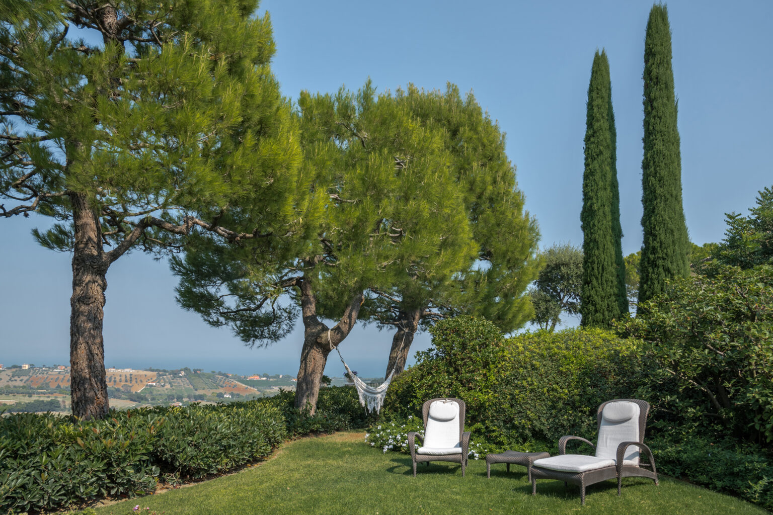 Serene outdoor garden scene featuring two lounge chairs on a lush green lawn, surrounded by tall cypress trees and panoramic views of the landscape. Ideal for relaxation and enjoying nature. | Only on SopranoVillas