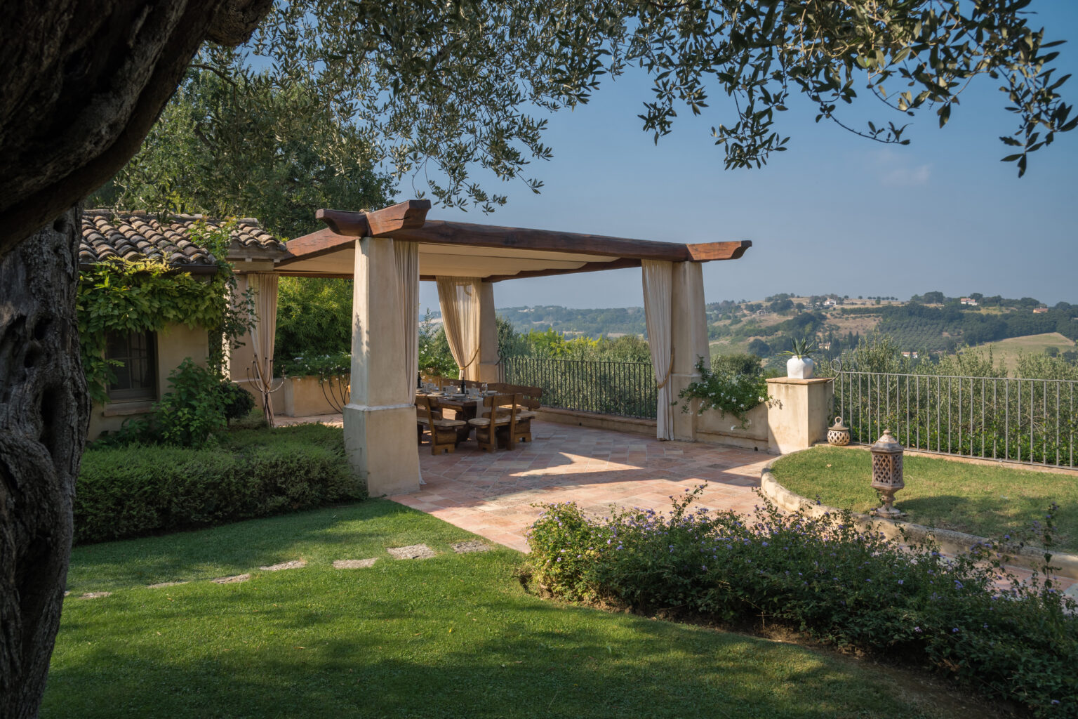 Outdoor patio with a pergola, dining table, and lush greenery, overlooking rolling hills and a serene landscape. Ideal for relaxation and entertaining in a beautiful garden setting. | Only on SopranoVillas