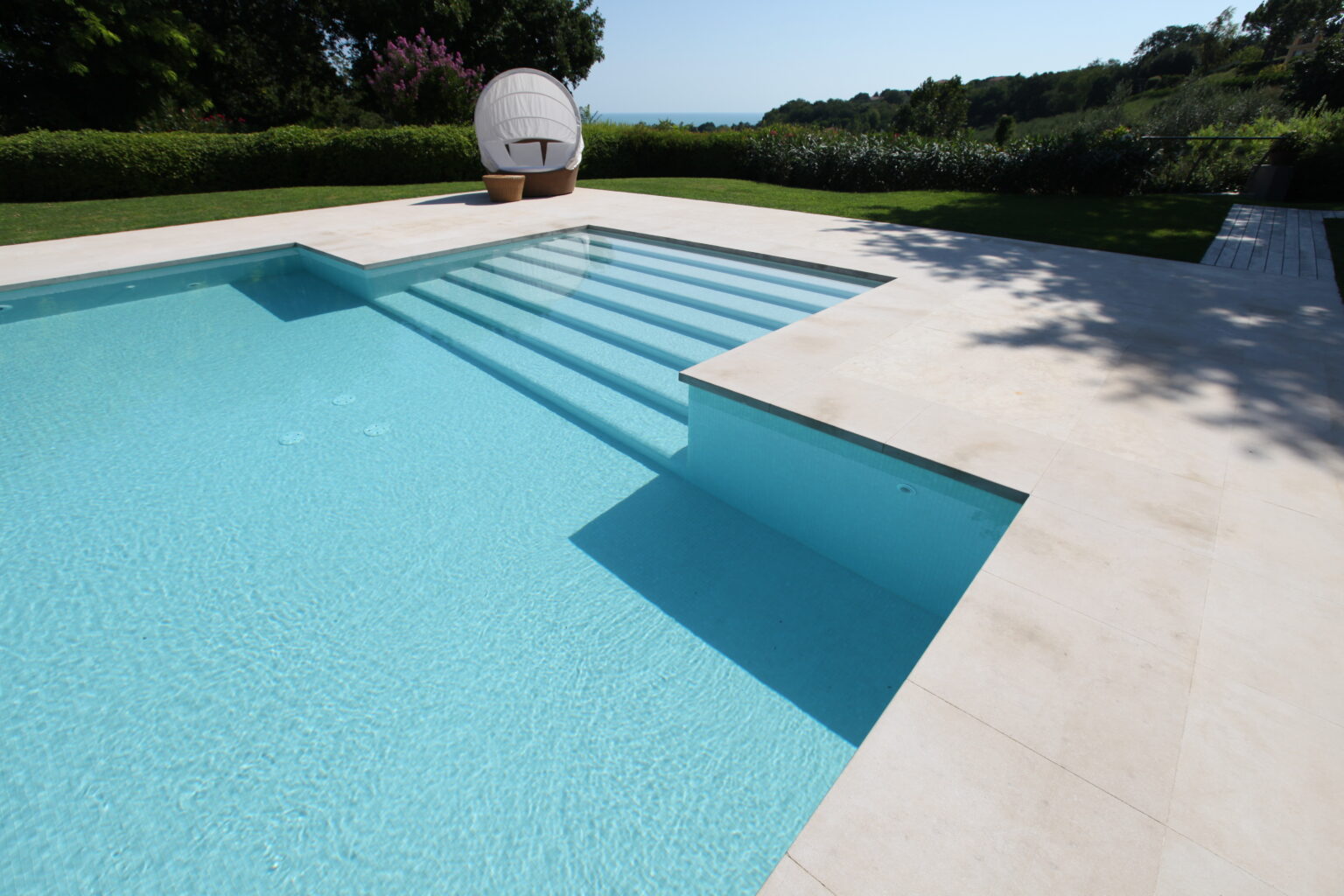 Modern swimming pool with clear blue water, featuring elegant steps, surrounded by lush greenery and a stylish lounging area. Ideal for outdoor relaxation and leisure. | Only on SopranoVillas