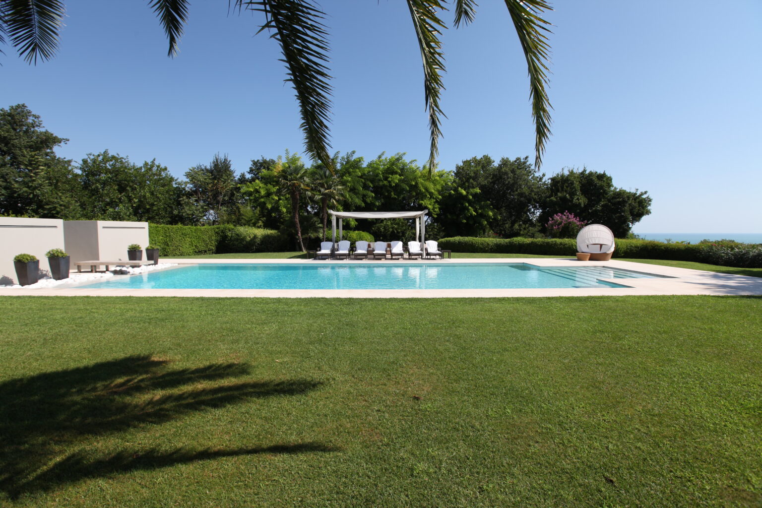 Luxurious outdoor swimming pool surrounded by lush green grass and palm trees, featuring comfortable lounge chairs and a shaded seating area, ideal for relaxation and leisure. | Only on SopranoVillas