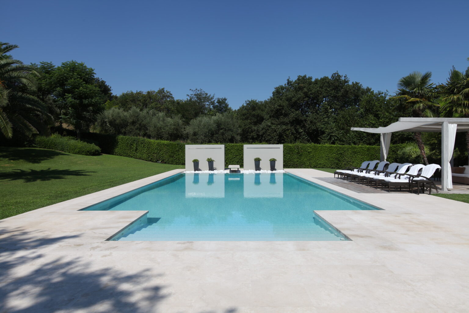 Modern outdoor swimming pool surrounded by lush greenery, featuring comfortable lounge chairs and a shaded seating area under a pergola. Perfect for relaxation and leisure in a sunny environment. | Only on SopranoVillas