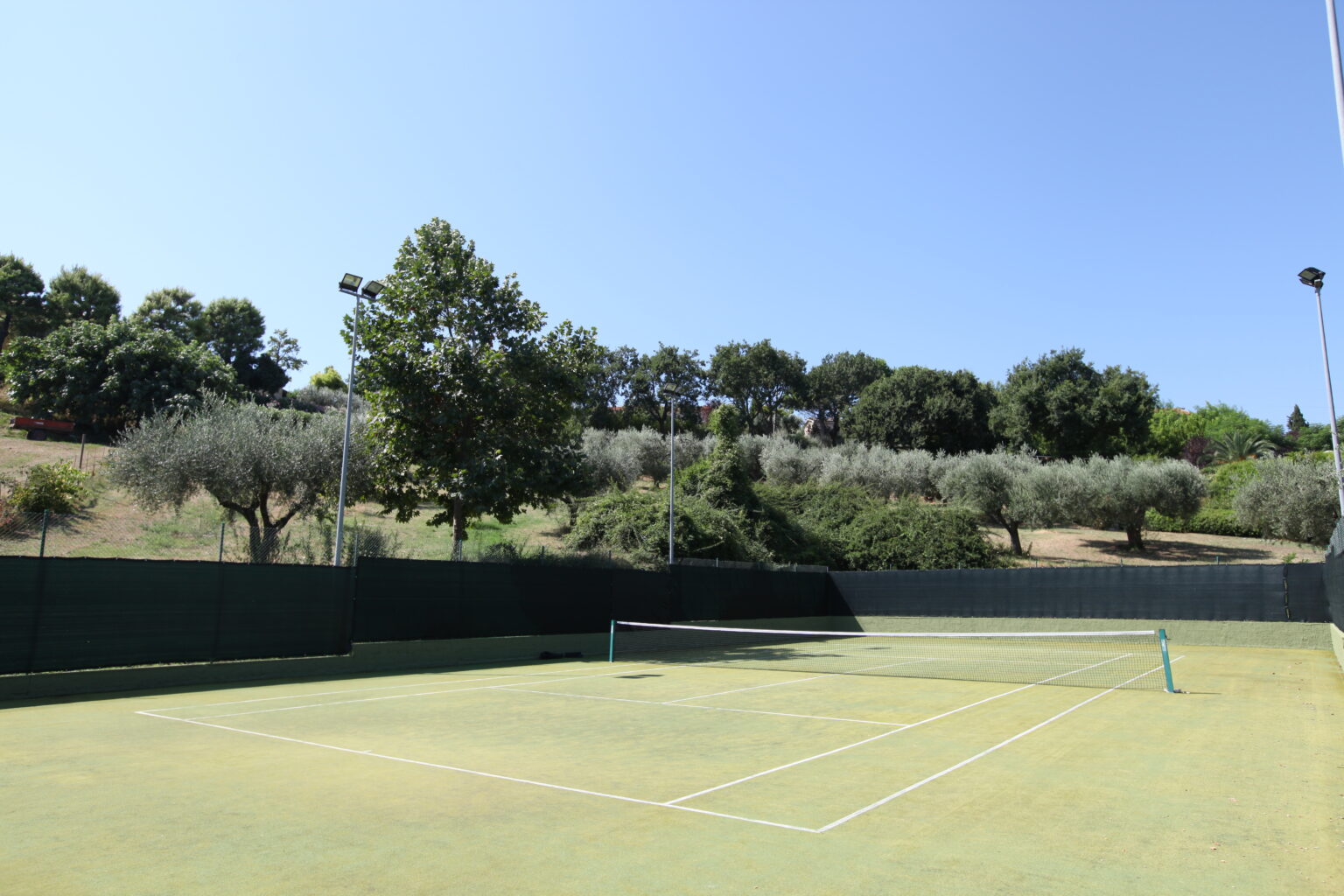 Tennis court surrounded by greenery under a clear blue sky, featuring a net, trees, and a hillside with olive trees in the background. | Only on SopranoVillas