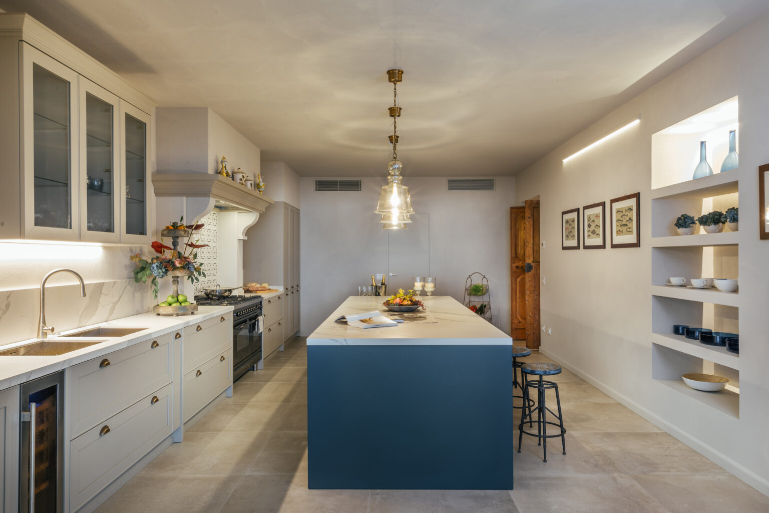 Modern kitchen design featuring a spacious island with a dark blue base, white cabinetry, pendant lighting, and open shelving displaying decorative items. The room is well-lit with natural light, showcasing a minimalist aesthetic and a functional layout. | Only on SopranoVillas