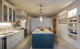 Modern kitchen design featuring a spacious island with a dark blue base, white cabinetry, pendant lighting, and open shelving displaying decorative items. The room is well-lit with natural light, showcasing a minimalist aesthetic and a functional layout.