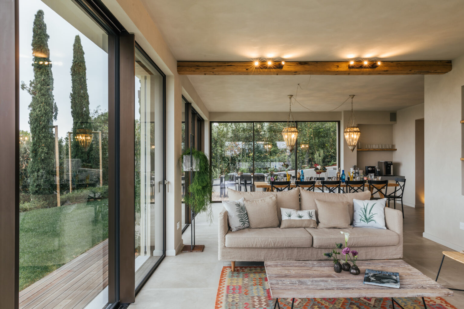 Modern living room interior featuring a beige sofa, decorative pillows, and a stylish coffee table, with large glass doors opening to a lush outdoor view. The space is illuminated by pendant lights and showcases a blend of natural and contemporary design elements. | Only on SopranoVillas