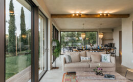 Modern living room interior featuring a beige sofa, decorative pillows, and a stylish coffee table, with large glass doors opening to a lush outdoor view. The space is illuminated by pendant lights and showcases a blend of natural and contemporary design elements.