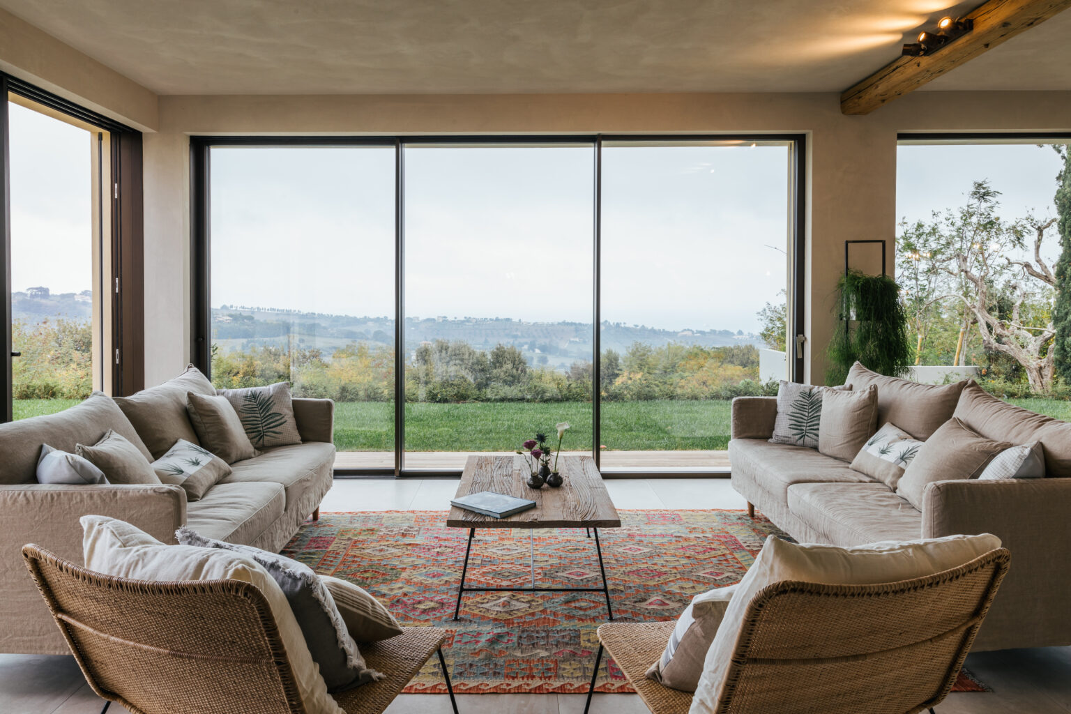 Modern living room with large windows offering scenic views, featuring cozy beige sofas, a wooden coffee table, and stylish armchairs. The space is decorated with a colorful area rug and indoor plants, creating a warm and inviting atmosphere. | Only on SopranoVillas