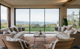 Modern living room with large windows offering scenic views, featuring cozy beige sofas, a wooden coffee table, and stylish armchairs. The space is decorated with a colorful area rug and indoor plants, creating a warm and inviting atmosphere.
