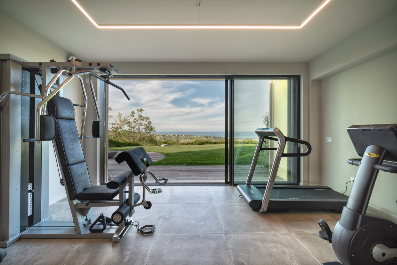 Modern home gym featuring a treadmill and multi-gym equipment, with large windows offering a scenic view of greenery and a bright, airy atmosphere. Ideal for fitness enthusiasts looking to exercise in a stylish environment. | Only on SopranoVillas