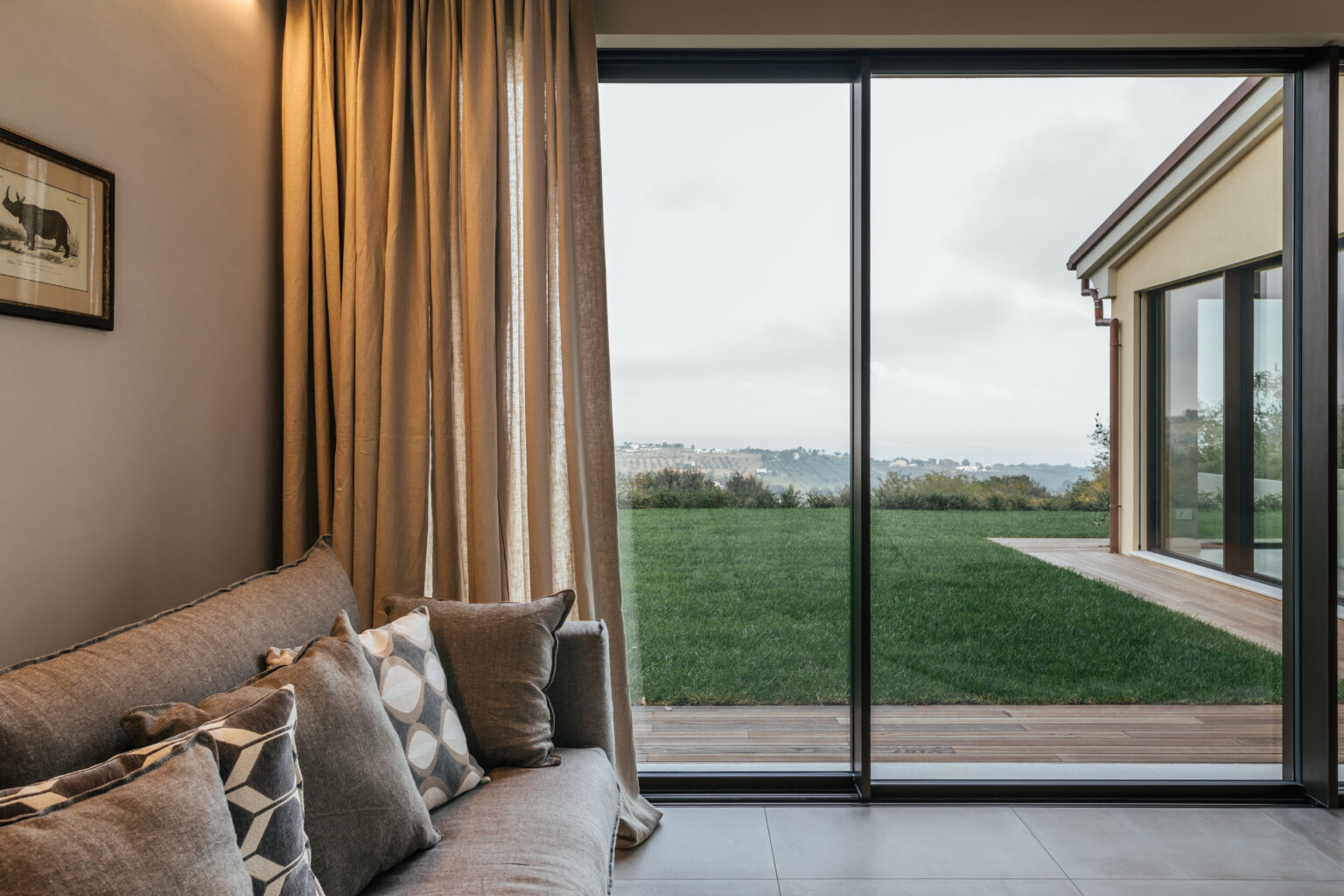 Cozy living room with a gray sofa and decorative pillows, featuring large glass sliding doors that open to a green outdoor space and scenic view. Soft, warm lighting enhances the inviting atmosphere. | Only on SopranoVillas