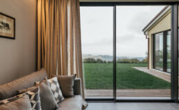 Cozy living room with a gray sofa and decorative pillows, featuring large glass sliding doors that open to a green outdoor space and scenic view. Soft, warm lighting enhances the inviting atmosphere.
