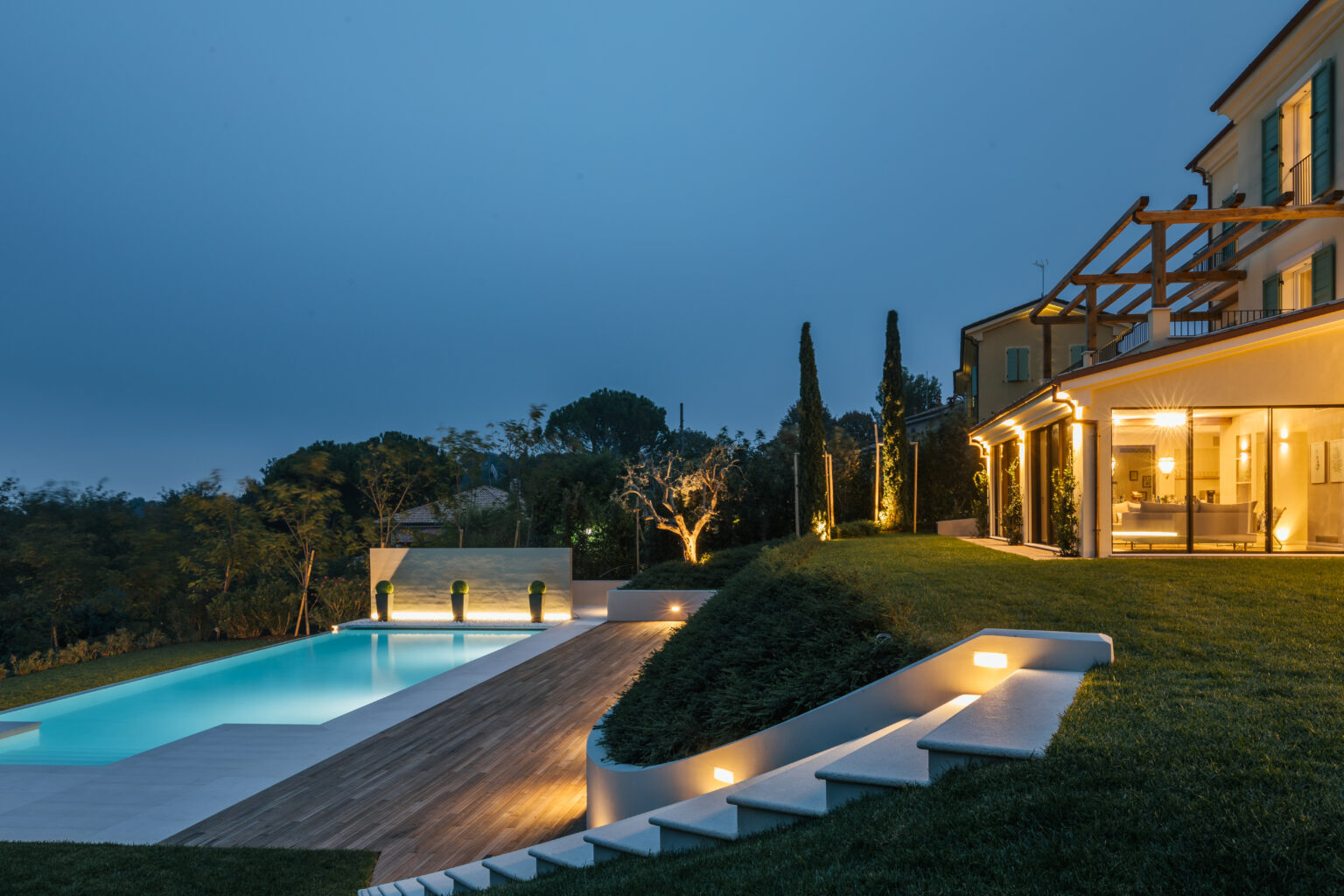 Modern villa exterior at dusk featuring a beautifully lit swimming pool, landscaped garden, and elegant architectural design, surrounded by lush greenery. | Only on SopranoVillas