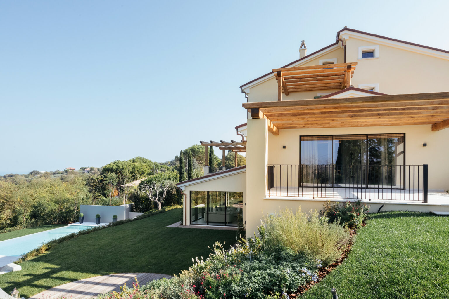 Modern villa with a spacious garden and swimming pool, featuring large windows and wooden pergola, surrounded by lush greenery and a clear blue sky. Ideal for luxury living and outdoor relaxation. | Only on SopranoVillas